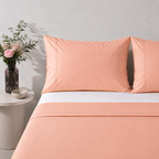 A neatly made bed with peach-colored 250TC organic cotton sheet and two matching pillowcases, styled in a minimalist bedroom setting with a small side table and flowers. with 250TC organic fitted sheet
