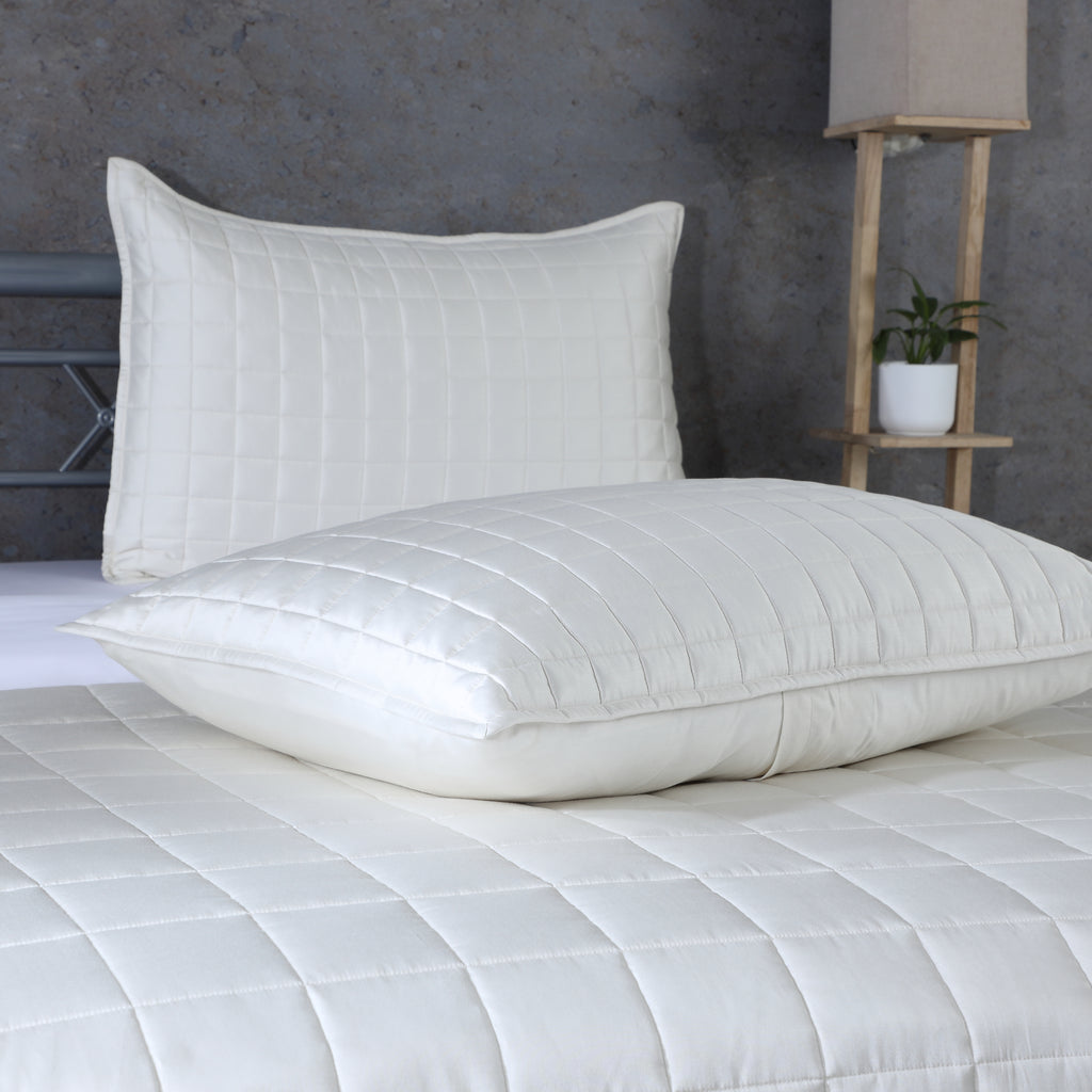 Two ivory pillowcases from Bamboo Cotton Bedspread Set stacked on bed showing quilted square stitching detail, styled with concrete wall and shelf, Whites of London NZ 