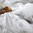 White bedding set with floral embroidery, a cup of tea, and small plant on a wooden tray.