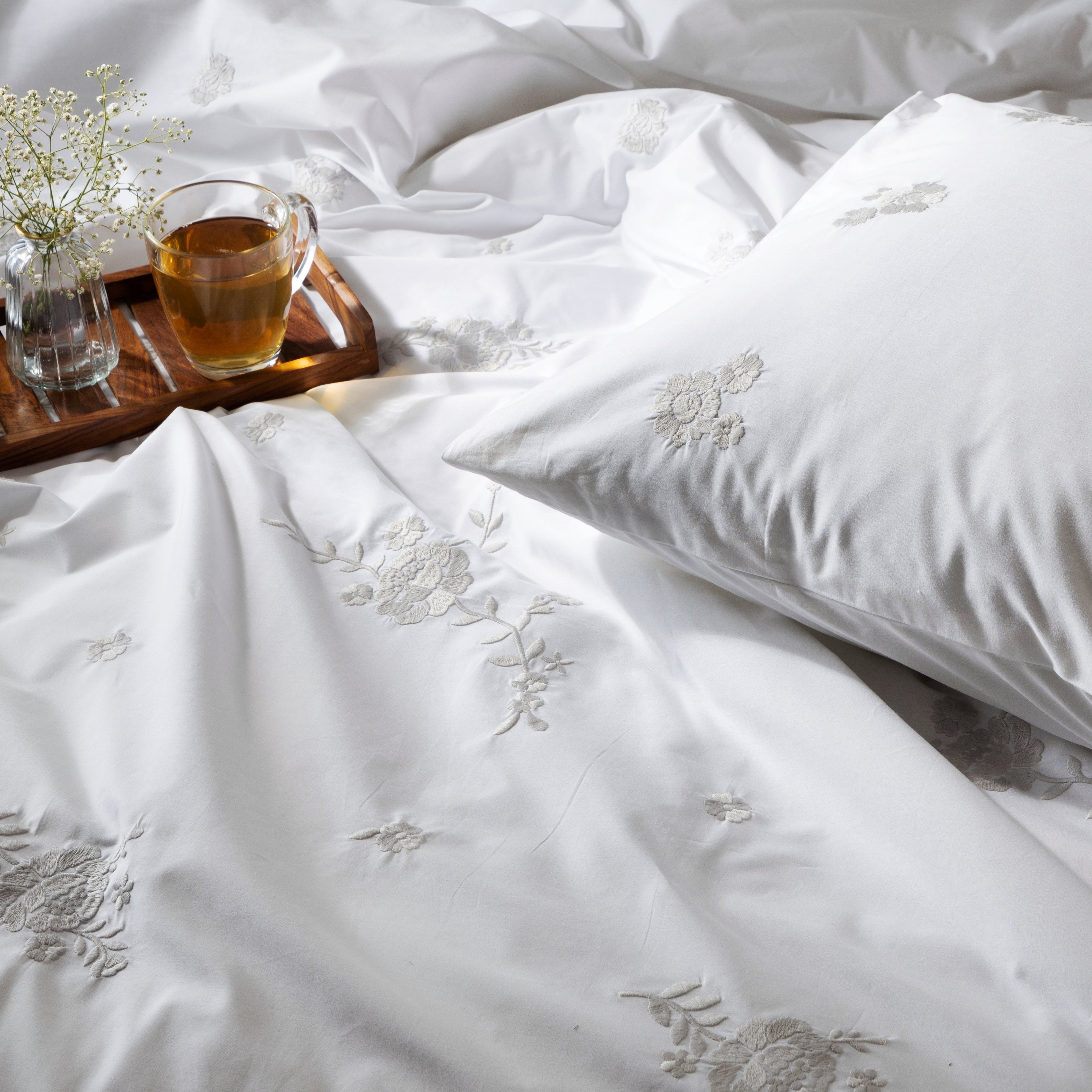 White bedding set with floral embroidery, a cup of tea, and small plant on a wooden tray.